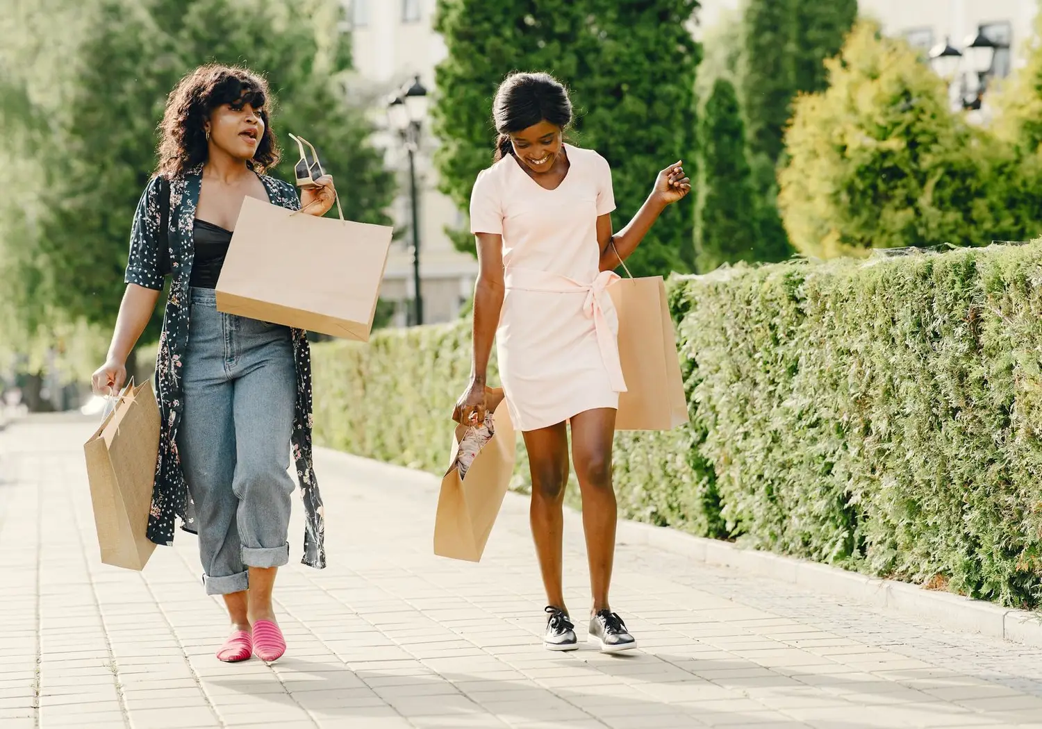 Duas mulheres caminham numa rua arborizada com sacos de compras.